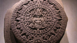 The image displays the Aztec calendar stone, also known as the Sun Stone, featuring intricate carvings. The calendar is a large circular stone with detailed mythological and astronomical engravings that played a significant role in the Aztec civilization. The central face of the sun god Tonatiuh appears at the center, surrounded by elaborate designs that represent different periods of time and the cosmos according to Aztec beliefs. The stone is showcased against a plain backdrop, emphasizing its historical and cultural significance.