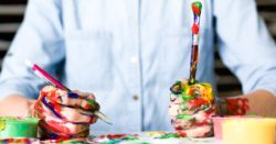 person holding click pen and a bursh with hands and table all covered in splashes of colorful paint