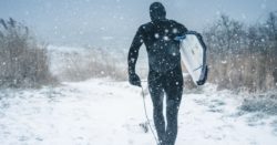 A man in a wet suit walking in the snow with the surfing board
